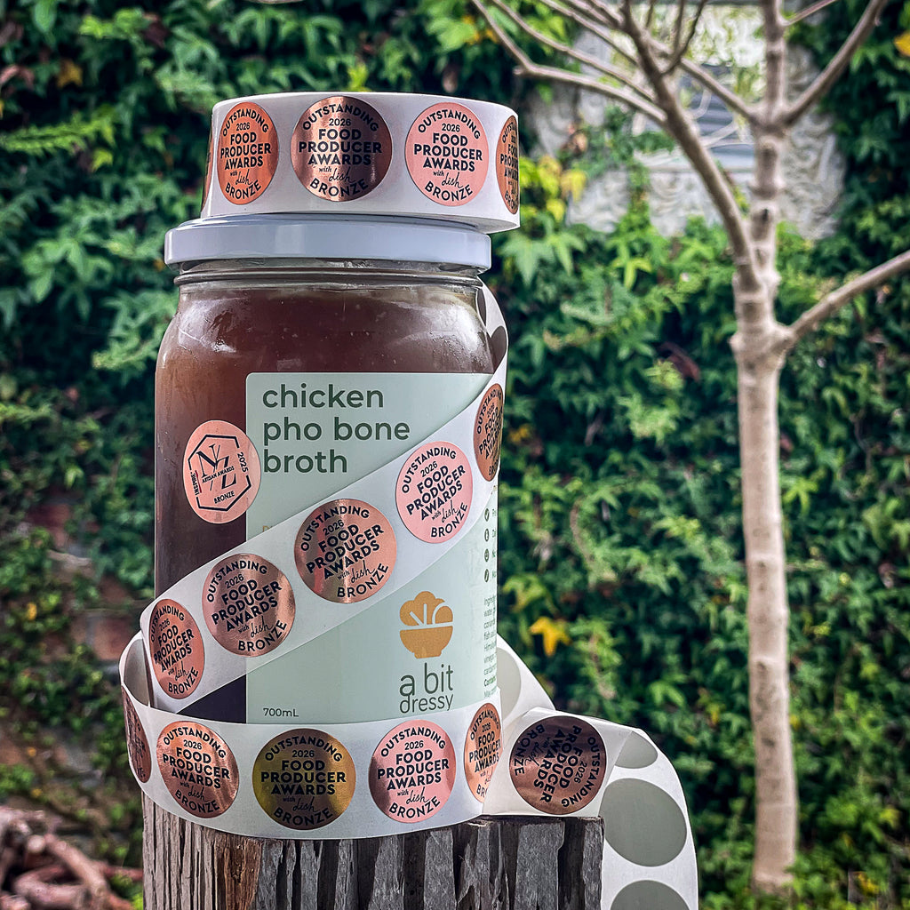 A glass jar of chicken pho bone broth by 'a bit dressy' sits on a rustic wooden stump with a lush, blurred green garden background.