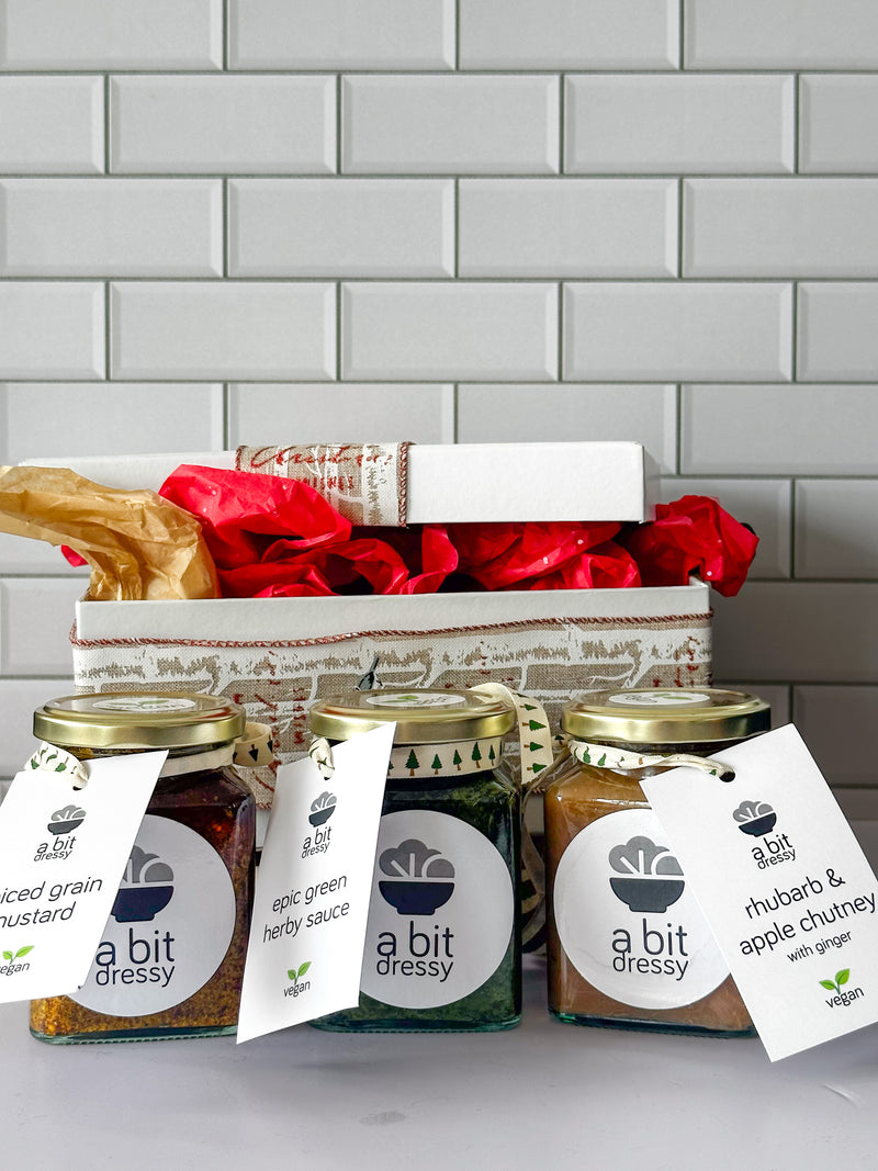 The Summer Condiment Bundle Christmas gift box, featuring three 270ml bottles of Spiced Grain Mustard, Epic Green Herby Sauce, and Lara's Glam Ham Glaze, along with a recipe tea towel, offering a delightful assortment of condiments and a practical kitchen accessory.