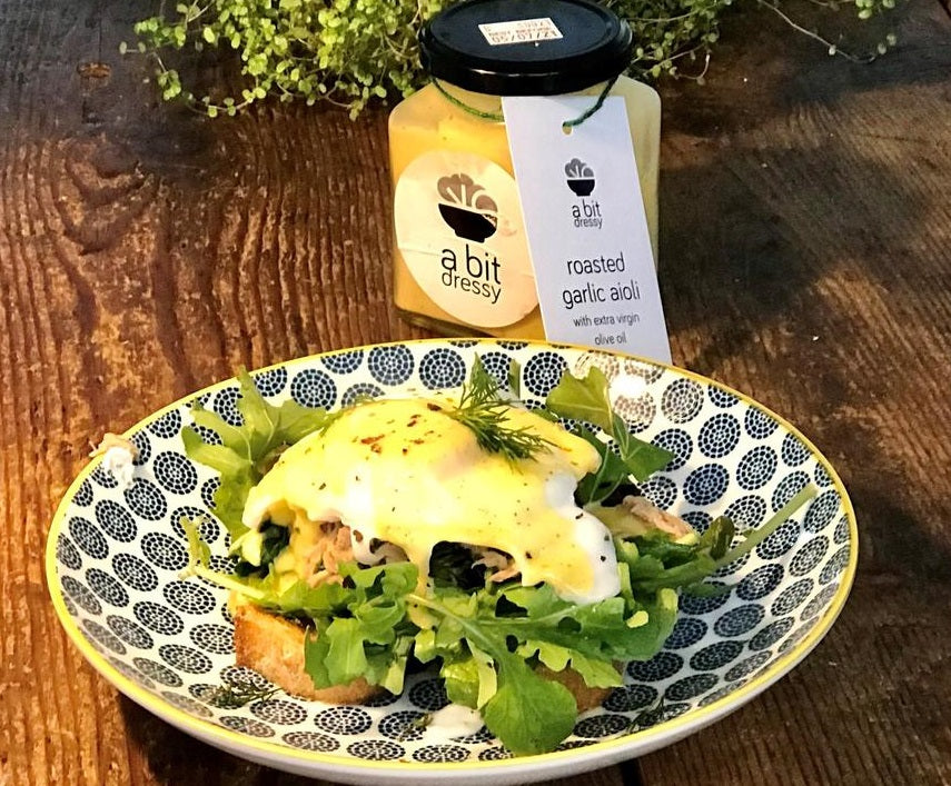 A jar of roasted garlic aioli with extra virgin olive oil drizzled generously over a plate of Eggs Benedict, adding a creamy and savory touch to the dish