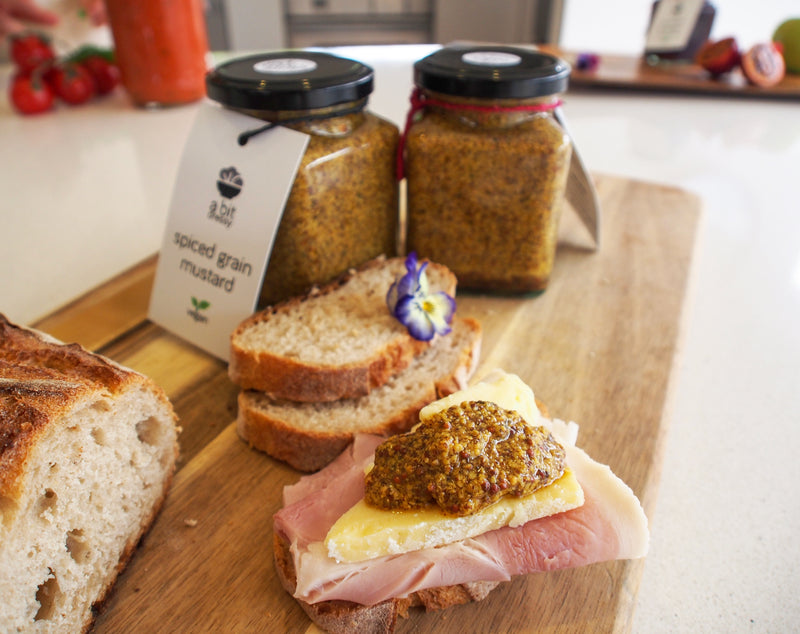  jar of spiced grain mustard next to a sandwich, with the mustard spread inside, adding a zesty and textured condiment to the sandwich.