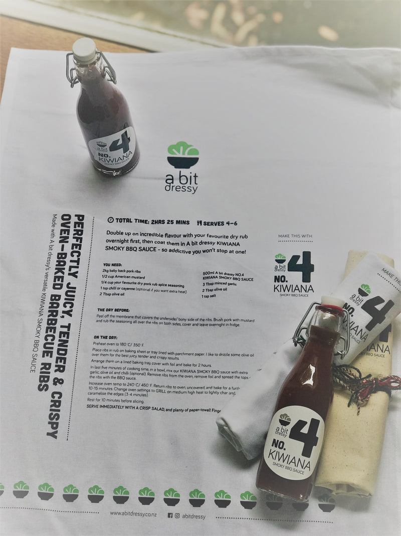A white tea towel displaying a BBQ ribs recipe that includes BBQ sauce from A Bit Dressy, positioned next to the BBQ sauce bottle, providing a helpful cooking guide and showcasing the condiment.